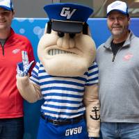 Two alumni, wearing the Red Wings GVSU hat, pose with Louie the Laker at the Detroit Red Wings GVSU Night